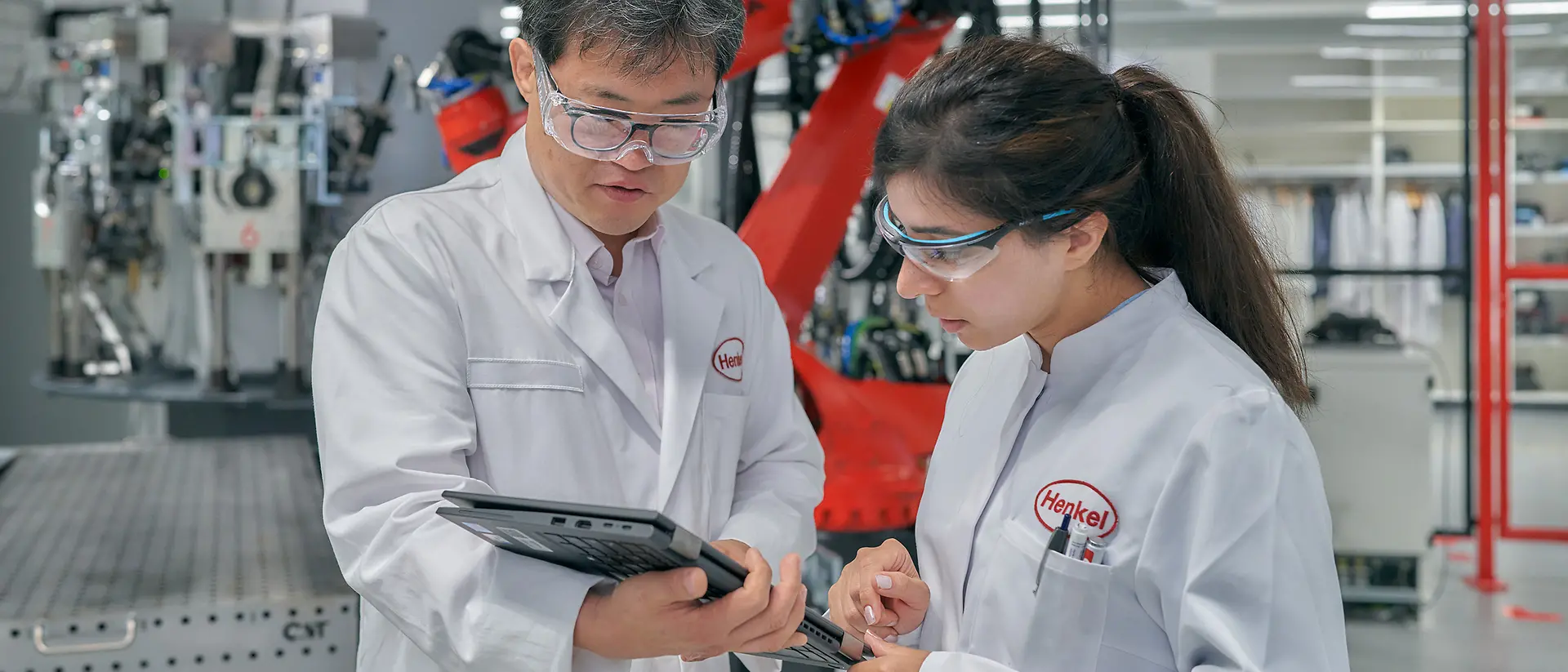A male and a female engineers wearing white, Henkel-branded coats look at a tablet. They are inside a factory with Henkel-branded machinery.