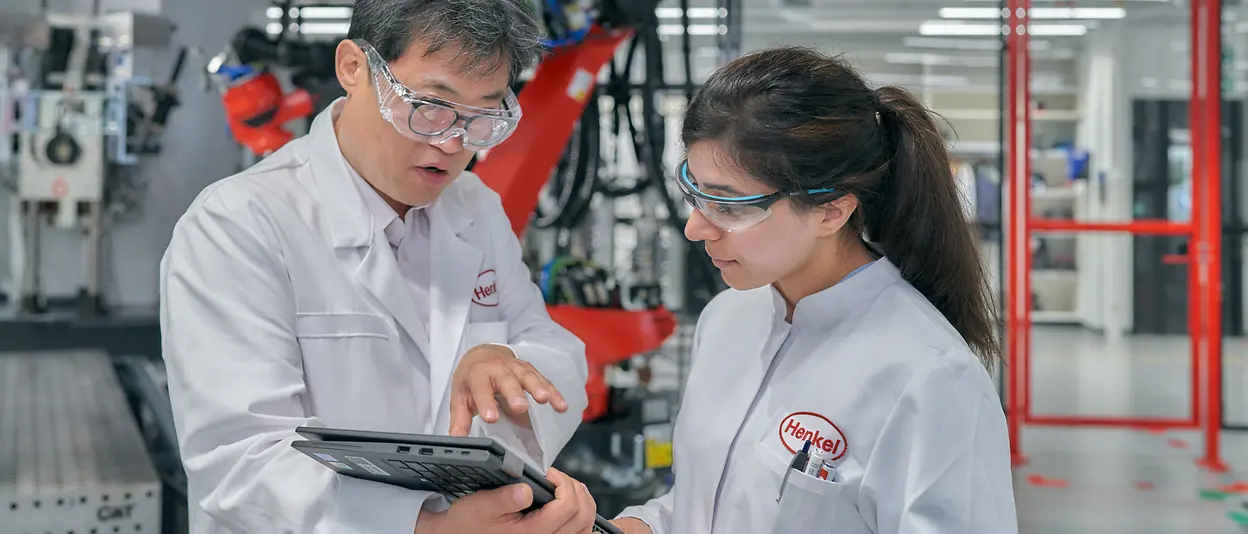 A male and a female engineers wearing white, Henkel-branded coats look at a tablet while talking to each other. They are on a factory floor.