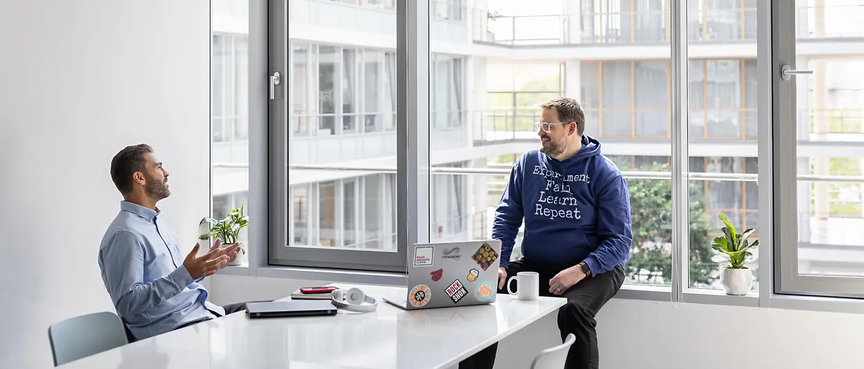 Two young men in a light office space talking in a relaxed atmosphere.