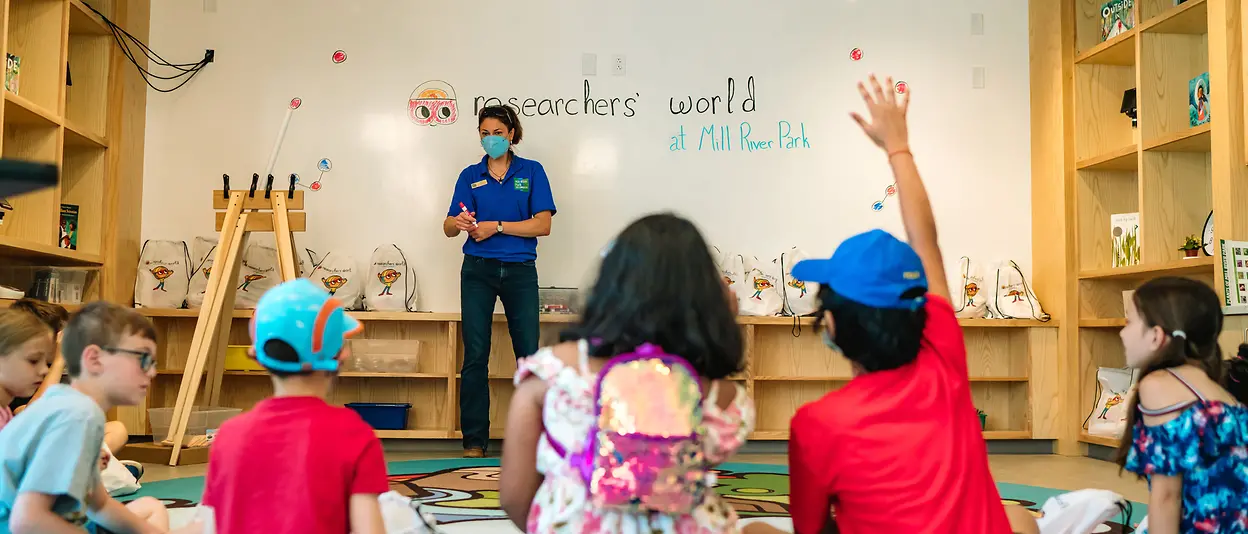 Woman wearing mask addresses children seated on classroom floor