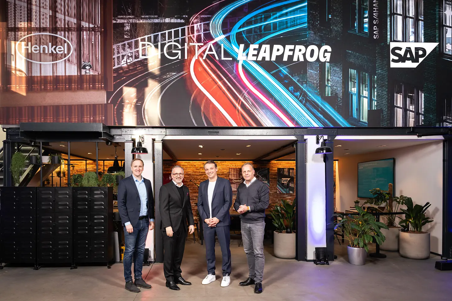 Four men standing in front a branded wall showing the Henkel and SAP logo. 