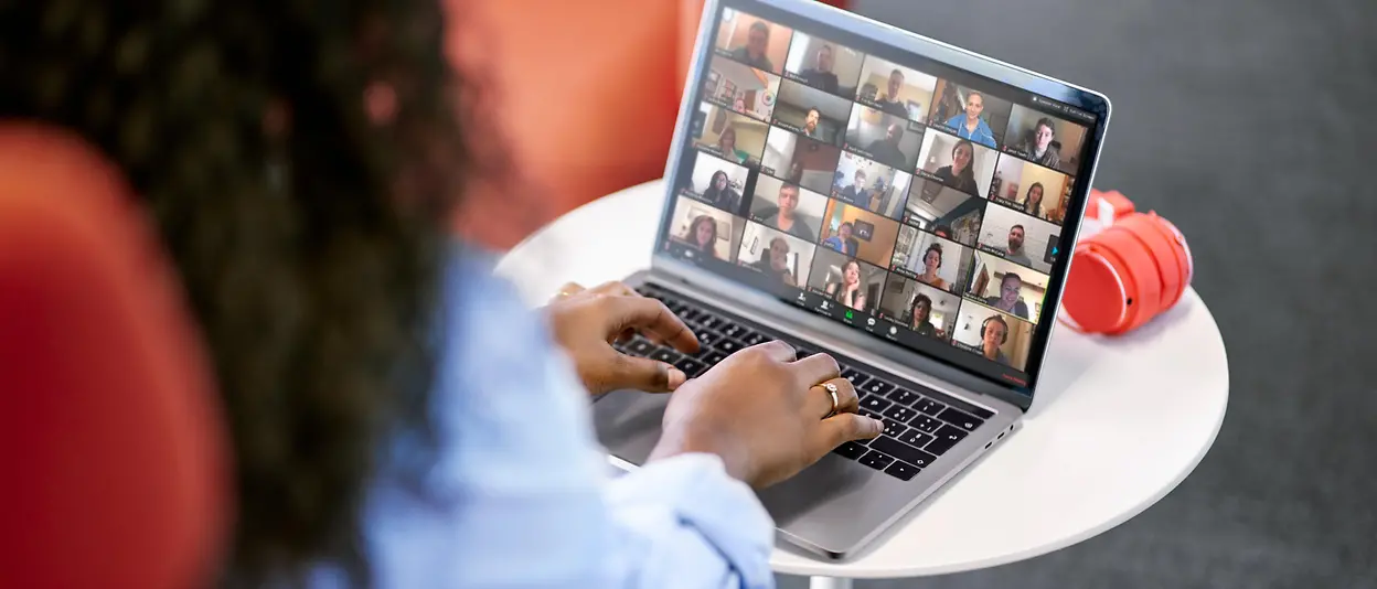 An employee sits at a table and participates in a digital gathering with her colleagues on her laptop.