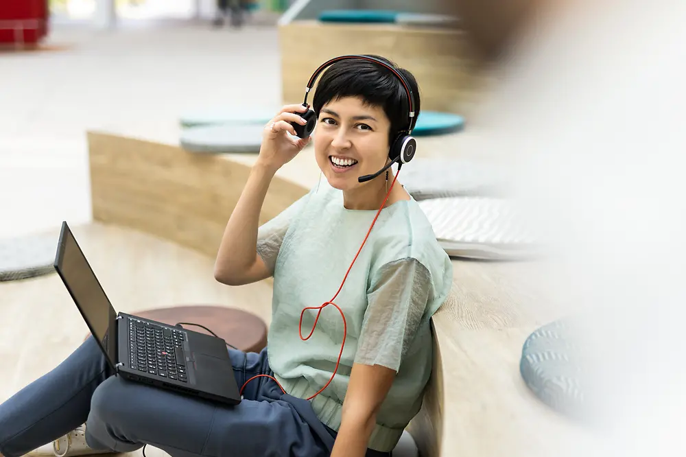 An employee has her laptop on her lap and wears a headset while smiling into the camera. 