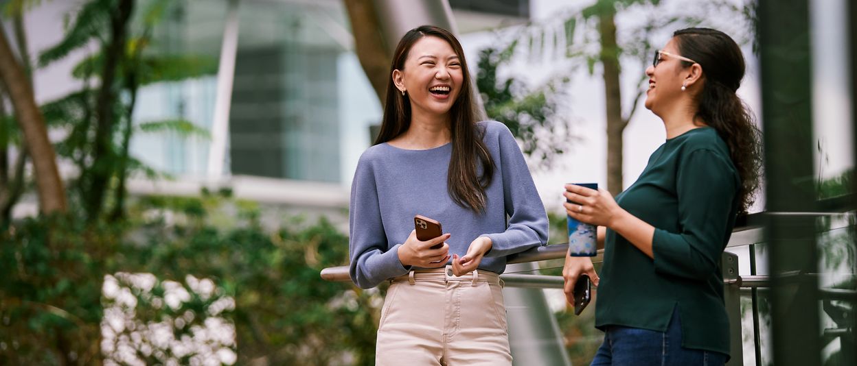 Two women stand next to each other laughing with their mobile phones in their hands.