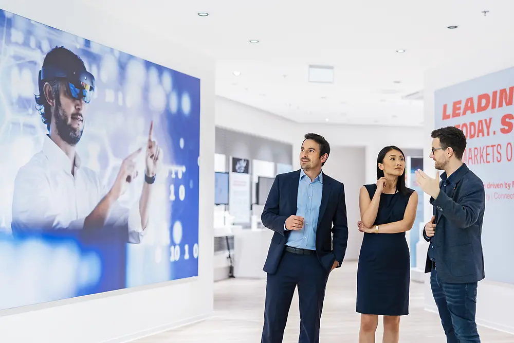 Two men and a woman are stadning and talking in a white room. One of the men is staring at a display showing a man wearing a VR headset.