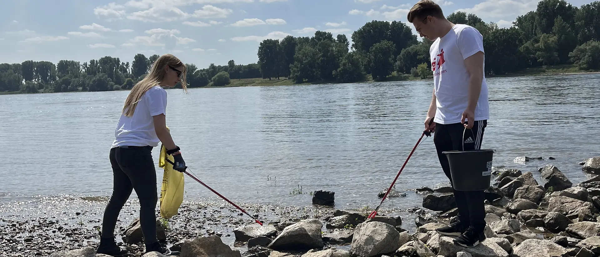 Two volunteers collect thrash with waste pickers during a rubbish collection event on a riverbank.