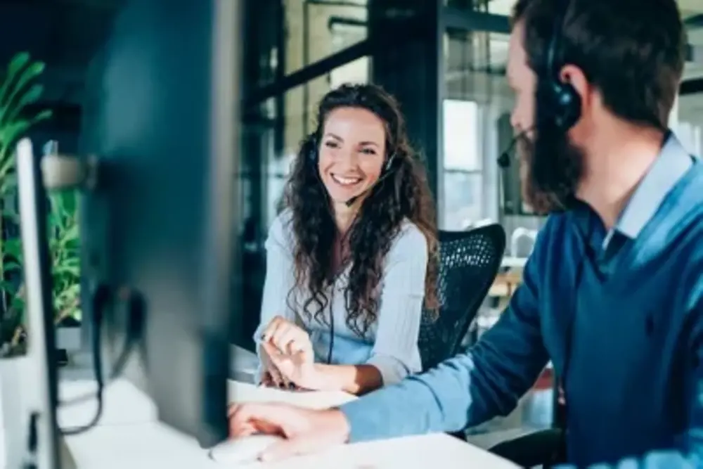 A man and a woman (boht with headsets) smile at each other while sitting in front of a computer.