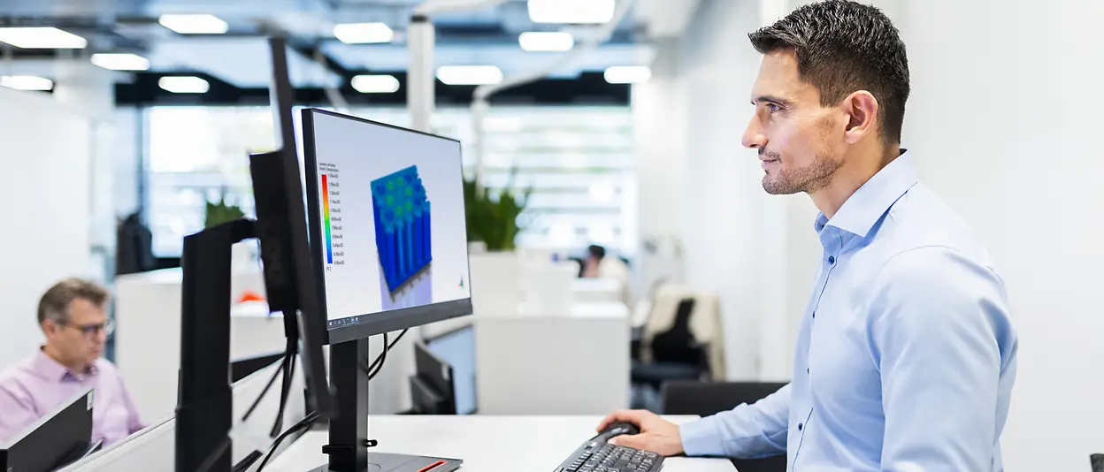 A man in a blue shirt is working at a computer. A 3D model can be seen on the screen. Other people working in the same office can be seen in the background.