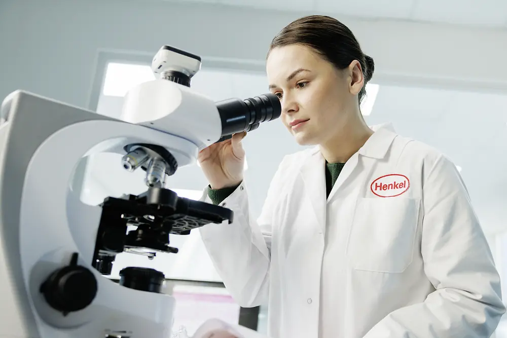 A woman in a lab coat with the Henkel logo looks through a microscope.