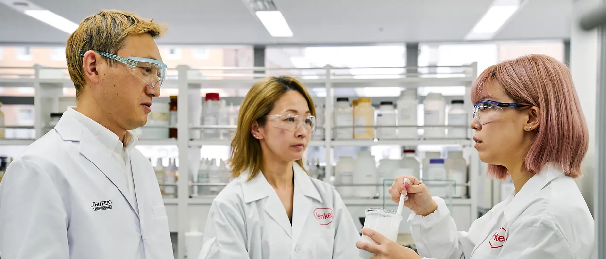 Three experts in white lab coats and protective goggles are standing in the lab talking about a formulation in a jar.