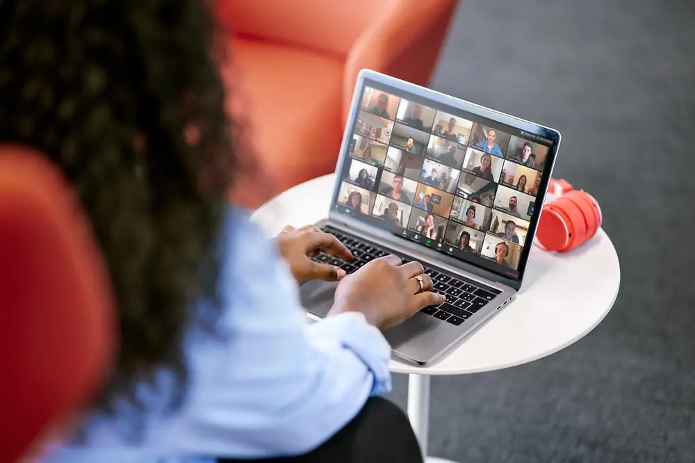 A woman with long brown hair and her back to the camera is sitting at a table with her laptop that displays a virtual meeting with colleagues.