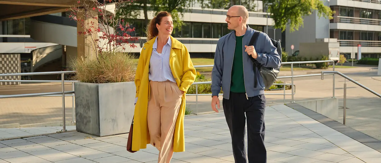 A woman in a yellow coat and a male colleague walk outside towards a building at Henkel.