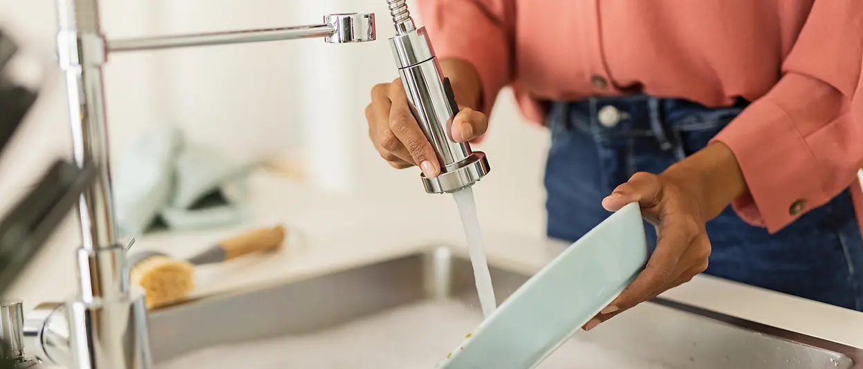 A woman standing at her kitchen sink cleaning dishes under running water