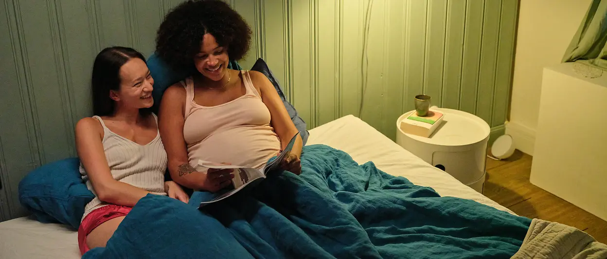 A female couple is lying together in bed, sharing a quiet, happy moment. One woman, visibly pregnant, smiles as she looks down at an open magazine she is holding. Her partner lies close beside her, gazing affectionately at her with a gentle smile. 