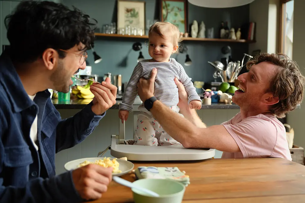 A baby girl is standing in a high chair at the dining table, supported by a smiling man who is gently holding her steady. Another man is sitting at the table, smiling warmly at her while taking a bite of food. The scene captures a joyful family moment, with both parents focused on the baby.