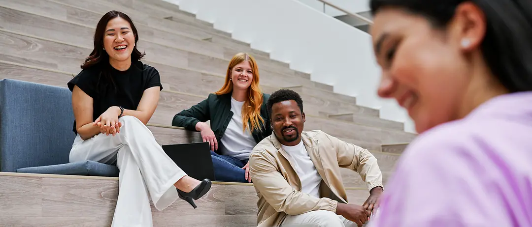 Three people sit on a staircase and watch as a fourth colleague presents something to them on a tablet.