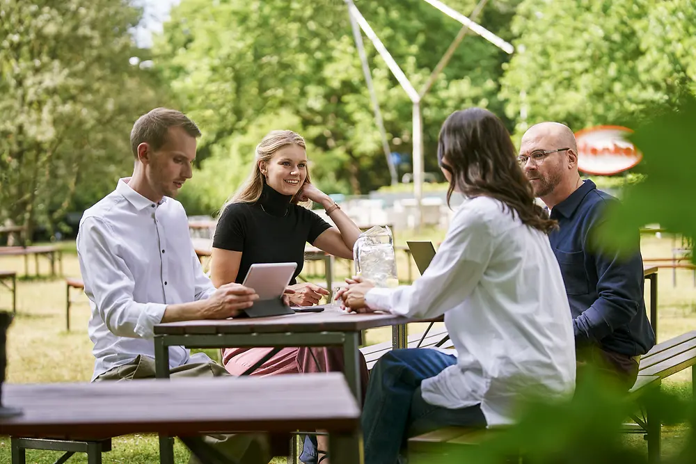 A group of four people sits with a laptop and a tablet at an outdoor table, engaged in conversation. Trees and a sunny setting are visible in the background. ​