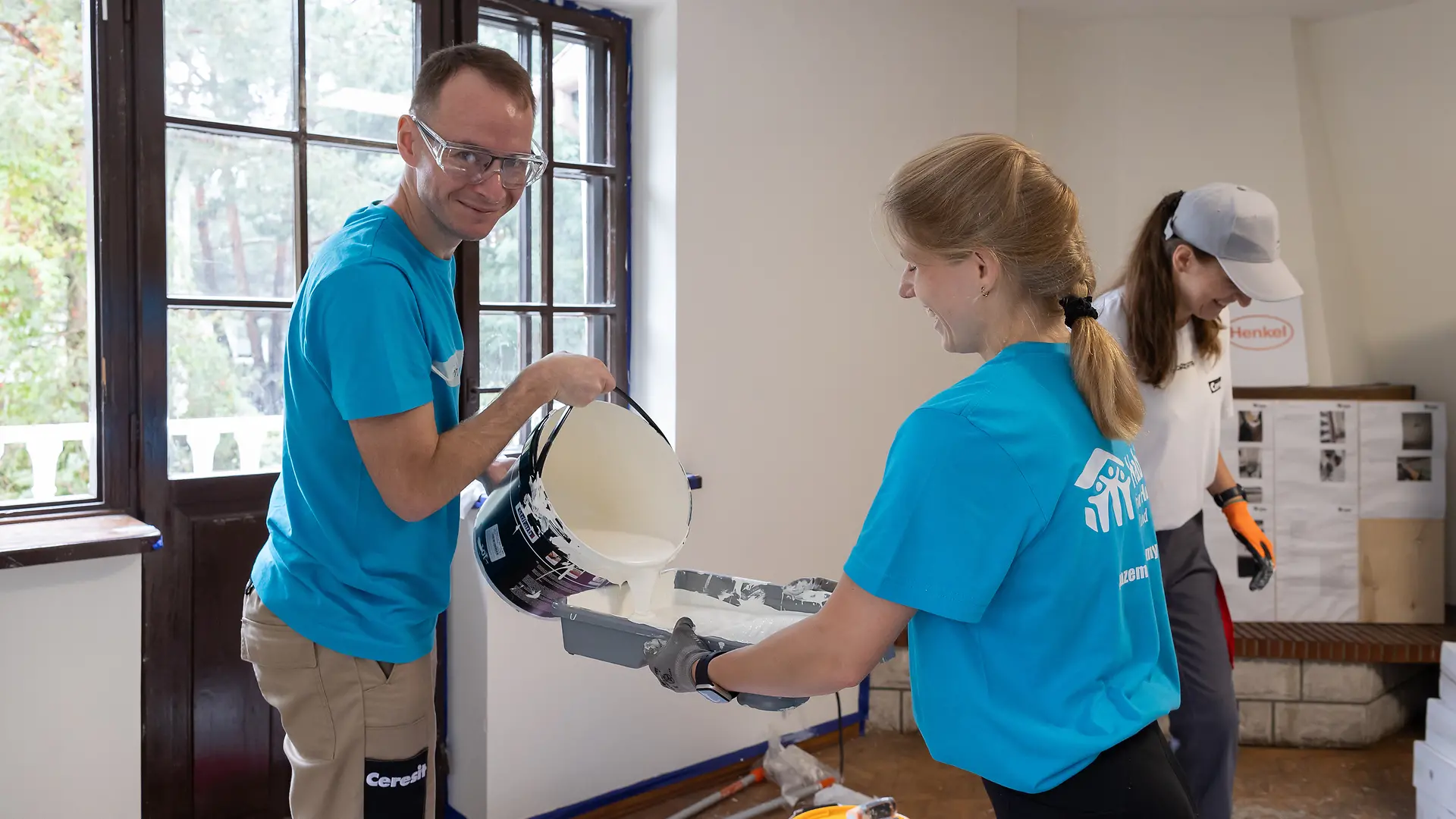 A Henkel employee pours white paint from a paint bucket into a paint tray held by a colleague.