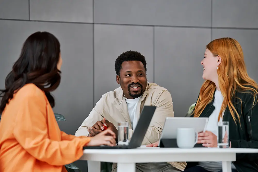 
Three people – two women and one man – are sitting at a table in a modern work environment, engaged in a lively conversation. Laptops and coffee cups are on the table.