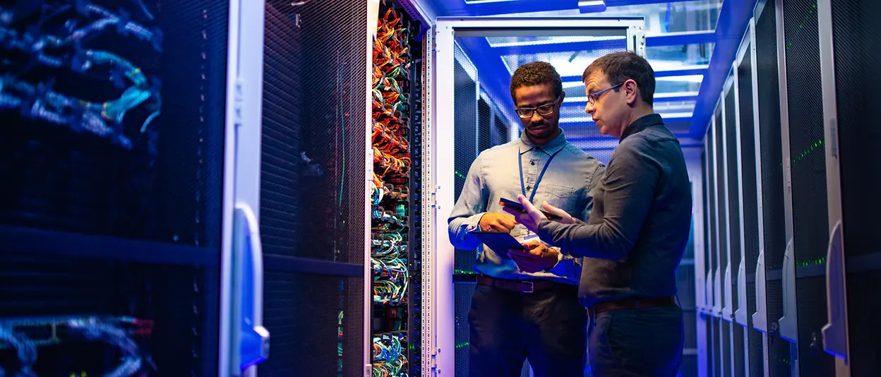 Two male engineers using devices in a server room