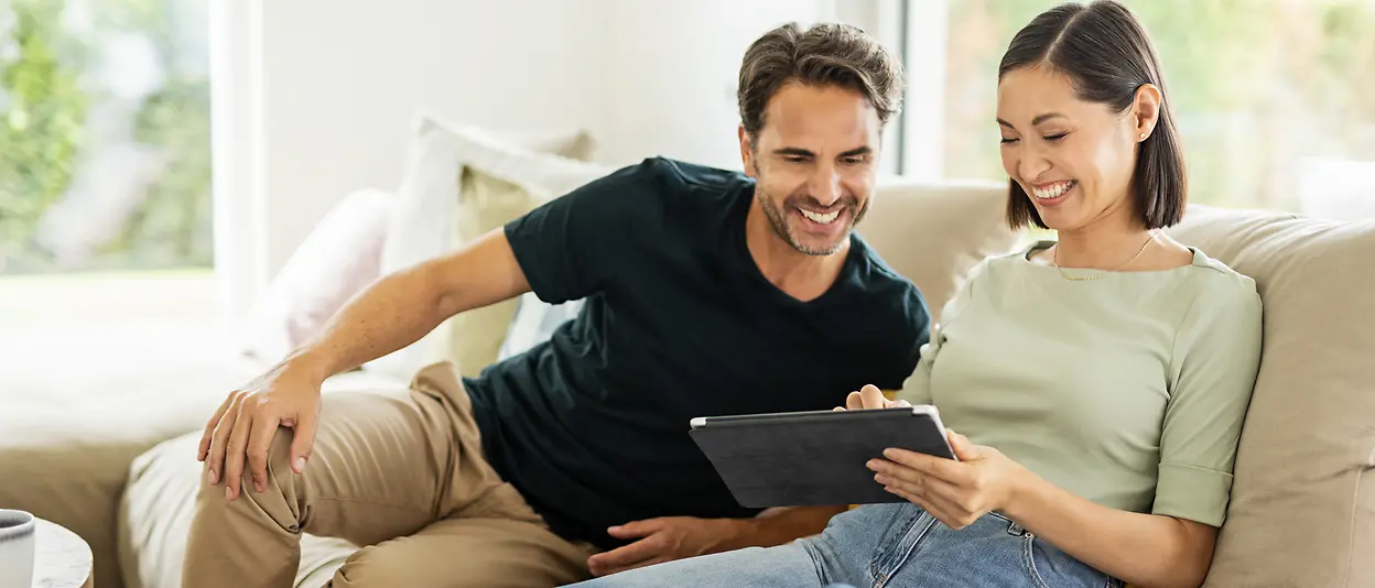 A man and a woman are sitting on their sofa at home looking at a tablet together.