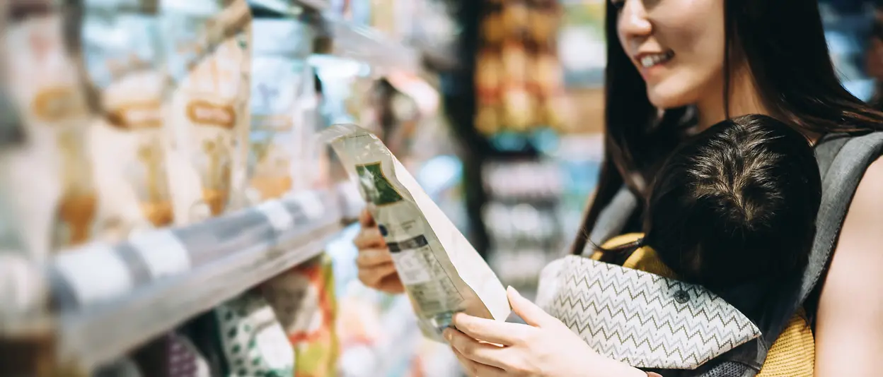 
A woman with her baby is standing in front of a shelf in a supermarket aisle, holding an item wrapped in a plastic pouch.